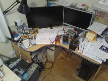 Nicholas Lee's second photo shows his desk with its PCs and a randomly strewn covering of schematics, test gear, oscilloscope, tools, and printed-circuit boards, as well as his trusty soldering iron.