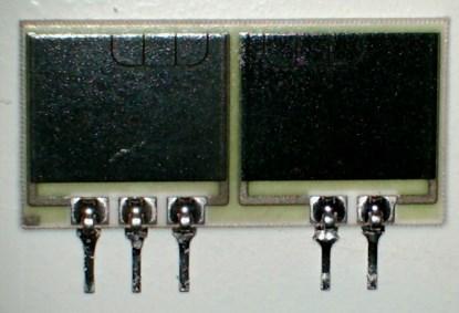 More printed film resistors on a ceramic substrate. This is a 2 x 600Ω resistor I obtained from a telephone PABX board (the middle connection on the group of three pins is not connected). I arranged the reflection of the light source so that the laser trimming on the left-hand one can clearly be seen. The whole assembly is just under an inch wide. These resistors are very accurate -- I saw no more than a 0.1Ω difference from 600Ω among six of them that I measured on my 4.5-digit multimeter.(Click here to see a larger image.)